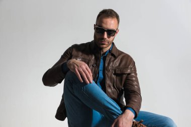 cool young guy with sunglasses holding knee up with hand above and posing in front of grey background in studio