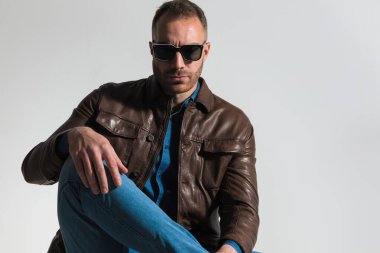 sexy casual man wearing blue denim shirt with brown leather jacket, holding knee up with hand on top on grey background in studio