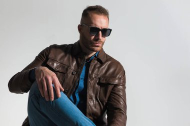 cool fashion man wearing sunglasses, holding knee up with hand above, looking away and posing on grey background in studio