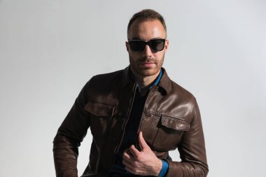 confident unshaved guy with sunglasses fixing brown leather jacket and posing on grey background in studio