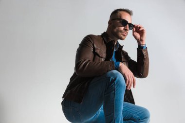 crouched young man holding arm on knee, fixing glasses and looking to side while posing in front of grey background in studio