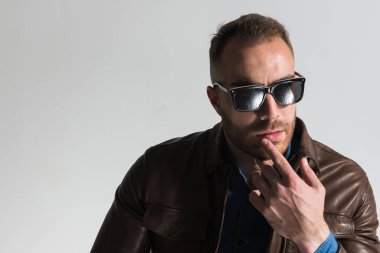 portrait of pensive casual guy wearing glasses, touching lips and thinking in front of grey background in studio
