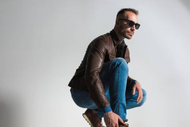 side view of casual fashion man in brown leather jacket crouching and looking to side in front of grey background in studio