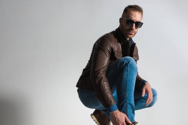 handsome bearded man in brown leather jacket crouching and posing in front of grey background in studio