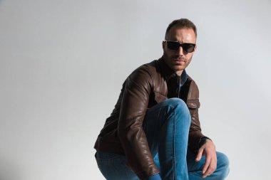 side view picture of sexy confident man in brown leather jacket crouching, wearing brown leather jacket and sunglasses and posing on grey background in studio