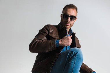 side view of handsome man adjusting and pulling brown leather jacket while crouching in front of grey background in studio