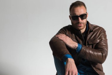 sexy bearded guy wearing sunglasses and brown leather jacket posing with arm on knee on grey background in studio