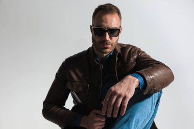 fashion man with beard posing with elbow on knee in a cool way in front of grey background in studio