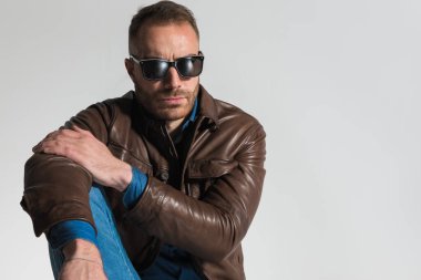 handsome casual man wearing brown leather jacket and posing with elbow on knee on grey background in studio