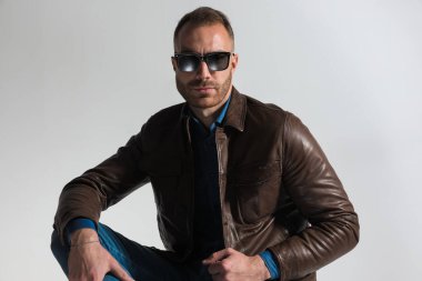 confident unshaved man wearing brown leather jacket and sunglasses and posing in front of grey background in studio