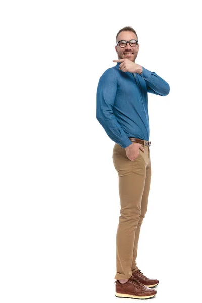 スタジオの白い背景に手でポケットに立って後ろに指を指すデニムシャツの幸せな若い男の側面図 — ストック写真