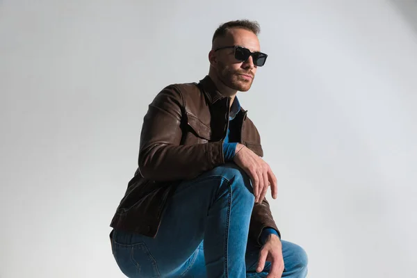 confident cool guy wearing brown leather jacket, crouching and looking away while posing on grey background in studio
