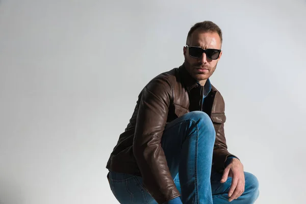 side view picture of sexy confident man in brown leather jacket crouching, wearing brown leather jacket and sunglasses and posing on grey background in studio