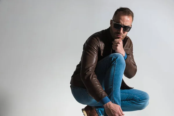 side view of cool fashion guy in brown leather jacket posing with fist on knee while crouching in front of grey background in studio