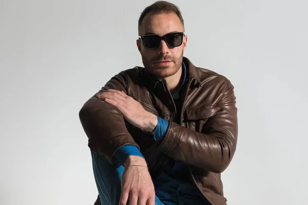 portrait of confident casual man wearing brown leather jacket and sunglasses, posing with elbow on knee in front of grey background in studio