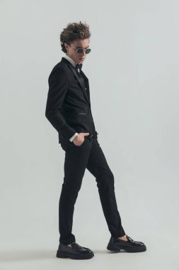 full body picture of elegant curly hair man with sunglasses holding hands in pockets, looking down and posing in front of grey background