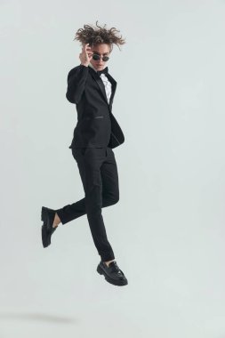 fashion elegant groom in tuxedo adjusting sunglasses while jumping in front of grey background in studio
