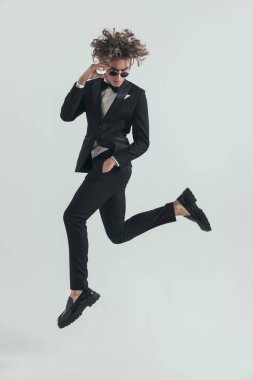 side view of sexy elegant man jumping in the air with hand in pockets and adjsuting sunglasses in front of grey background in studio