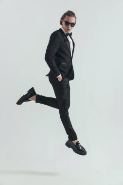 side view picture of elegant young man with sunglasses jumping in the air and posing with hands in pockets in front of grey background