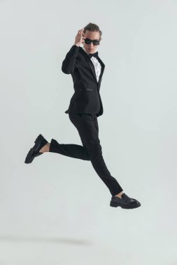 side view picture of cool elegant man jumping in the air and adjusting sunglasses in front of grey background in studio