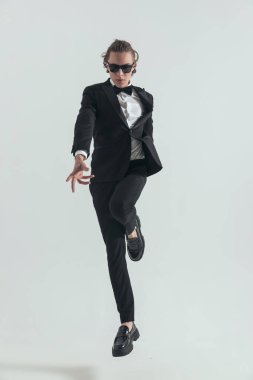full body picture of cool young groom in black tuxedo jumping up in the air and posing in front of grey background in studio