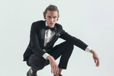picture of sexy elegant best man in black tuxedo crouching with elbows on knees and posing in a confident and serious manner on grey background