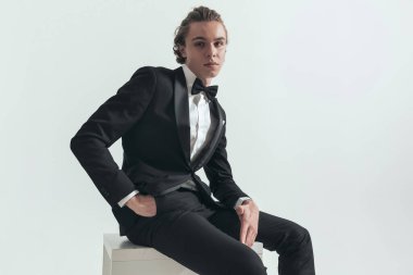 elegant young groom in tuxedo holding hand in pockets and posing while sitting on grey background
