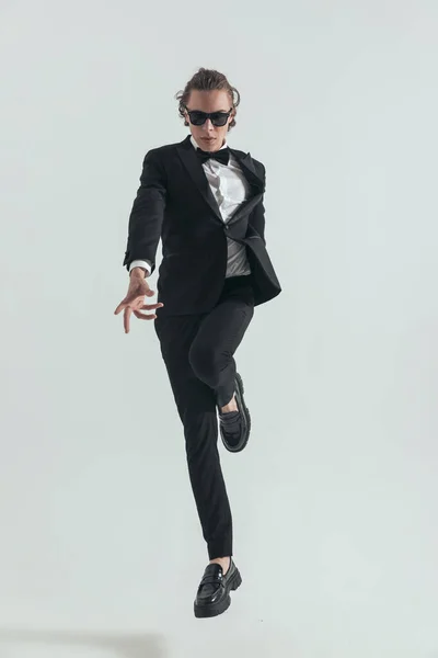 full body picture of cool young groom in black tuxedo jumping up in the air and posing in front of grey background in studio
