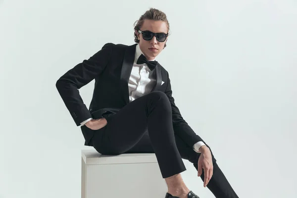 sexy best man in elegant tuxedo posing while sitting with hand in pockets in front of grey background in studio