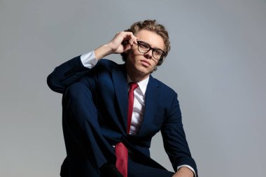 elegant businessman in suit looking away and fixing glasses while posing with elbow above knee in front of grey back