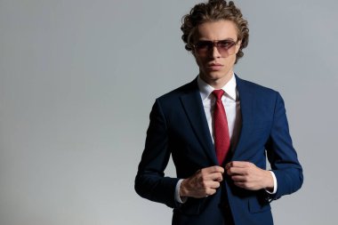 portrait of young man with long curly hair adjusting suit and posing in a cool way in front of grey background in studio