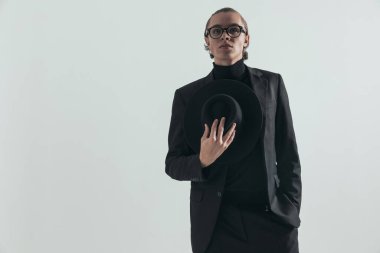 portrait of sexy fashion man in black suit holding hat in hand, looking up and posing with hand in pocket on grey background in studio