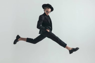 cool picture of confident fashion guy jumping in the air with hands in pockets while taking a huge step in front of grey background in studio