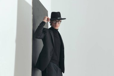 cool young guy with glasses adjusting hat while posing in front of black wall on grey background in studio