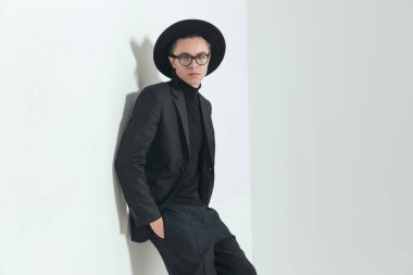 handsome young man with black suit holding hands in pockets and laying on a wall while posing on grey background in studio