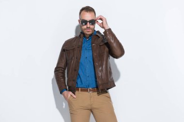 confident guy wearing blue denim shirt with brown leather jacket fixing glasses and posing in front of white background in studio