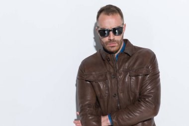 cool fashion guy in brown leather jacket with sunglasses posing in a confident way in front of white background in studio