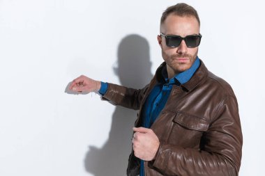 sexy confident man with sunglasses adjusting and pulling jacket and posing in front of white background in studio