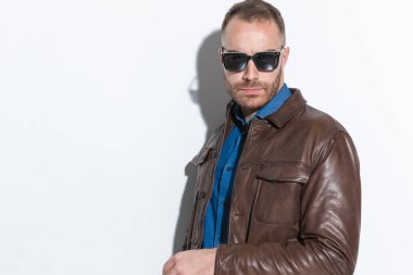 sexy young guy in brown leather jacket with sunglasses posing in front of white background in a cool and confident manner 