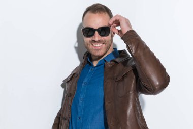 portrait of happy young man in brown leather jacket with jeans shirt smiling and adjusting sunglasses in front of white background in studio
