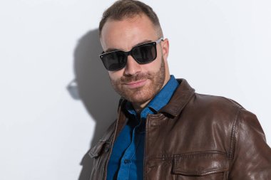 portrait of handsome casual man with sunglasses smiling and wearing brown leather jacket in front of white background in studio