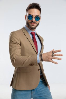 portrait of a attractive businessman posing with one open hand, standing, wearing sunglasses against gray studio background