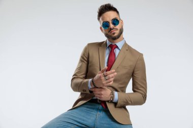 portrait of a handsome businessman fixing his bracelet, sitting, wearing sunglasses against gray studio background