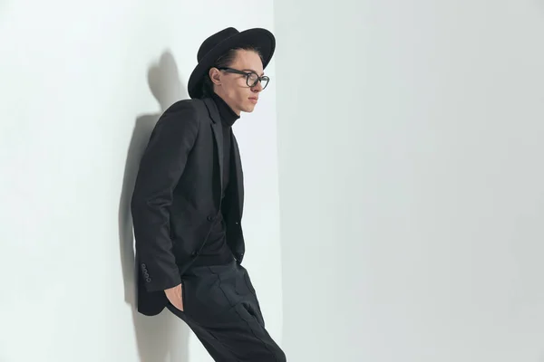 side view of fashion guy with hat and glasses, looking to side, laying on a wall and holding hands in pockets while posing on grey background