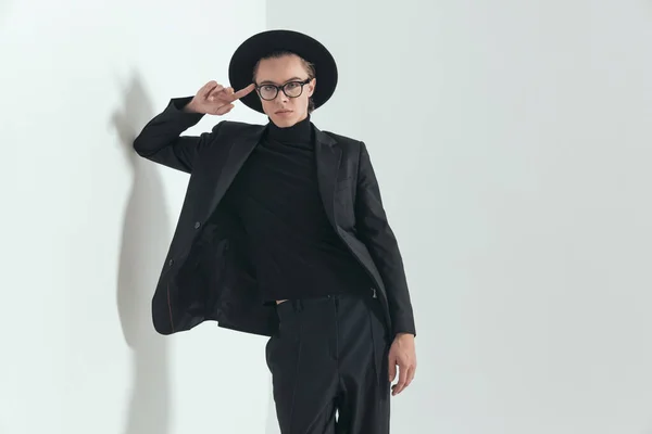 portrait of fashion guy wearing black suit with hat and glasses posing while laying with elbow on wall on grey background