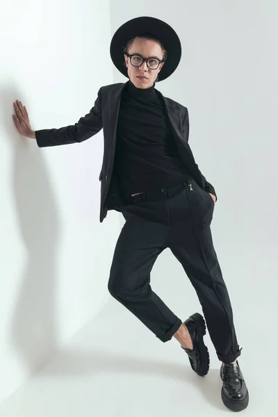 cool fashion guy with hat and glasses posing while laying on a wall and holding hand in pocket in front of grey background in studio