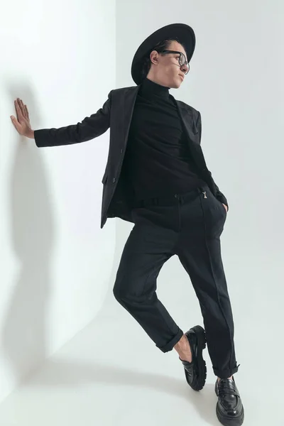 cool picture of fashion young man in blakc suit with hat holding hand in pocket and posing while laying on a wall on grey background in studio