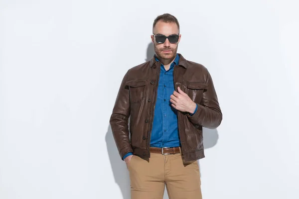 portrait of attractive young man with hand in pocket adjusting brown leather jacket on white background in studio