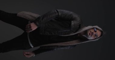vertical video of confident guy with hood rubbing palms and adjusting glasses in front of grey background in studio