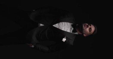 elegant man in black tuxedo with bowtie and handkerchief holding hands in pockets and looking to side while standing in spotlight